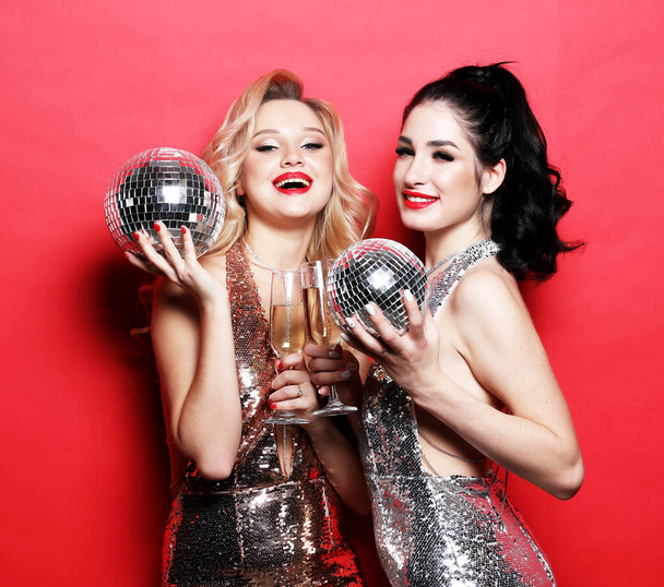 Δύο γοητευτικές γυναίκες, φιλενάδες σε πολυτελή glitter πούλιες φόρεμα και φωτεινό visage κρατώντας ντίσκο μπάλες και ένα ποτήρι κρασί, - Φωτογραφία, εικόνα