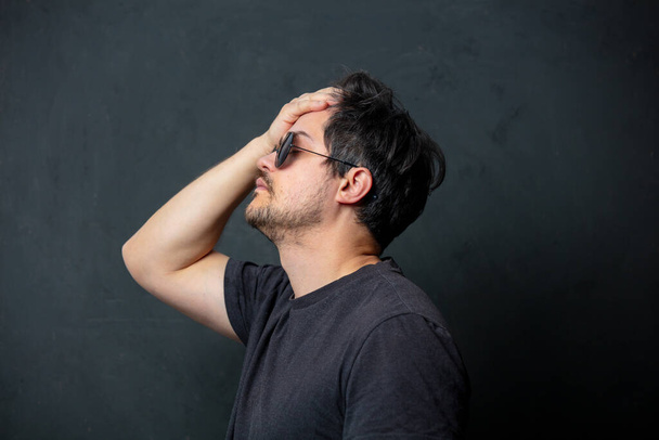 Tired brunet man in black t-shirt and sunglasses on dark background - Fotoğraf, Görsel