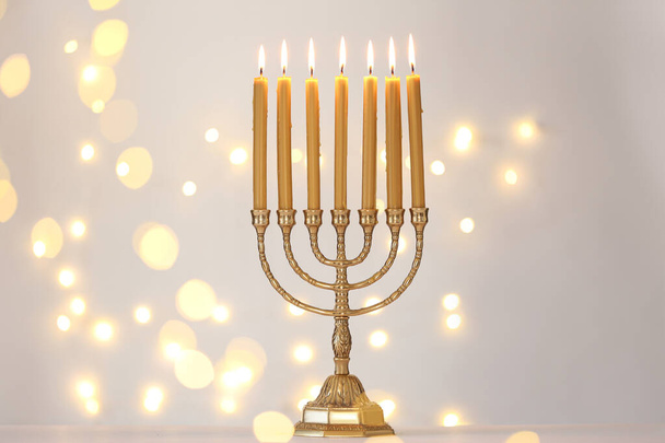 Golden menorah with burning candles against light grey background and blurred festive lights - Foto, afbeelding