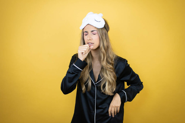 jonge mooie vrouw dragen slaap masker en pyjama over geïsoleerde gele achtergrond met haar hand naar haar mond hoesten - Foto, afbeelding