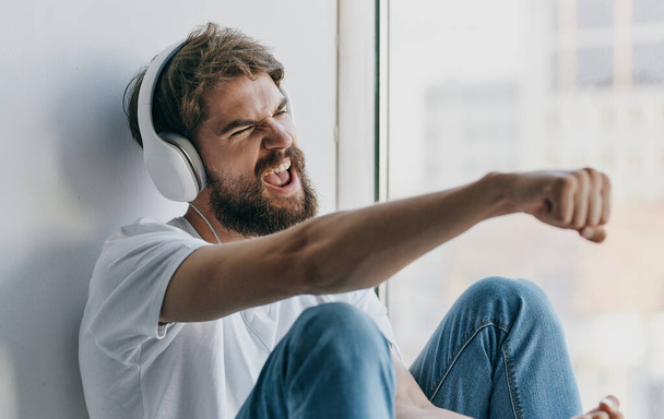 emotivní muž poslouchá hudbu na sluchátkách a gestikuluje rukama u okna - Fotografie, Obrázek