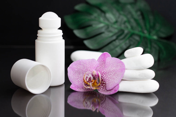 Beautiful lilac orchid flower and white roll-on deodorant with white stones and monstera leaves, on black background - Photo, Image