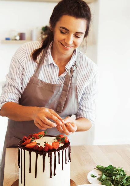 Young brunette female makes biscuit cake with chocolate and strawberries in the kitchen. - 写真・画像