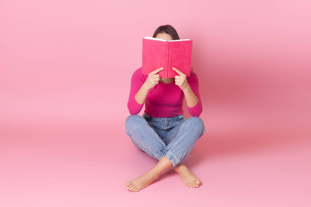 portrait of a young woman sitting cross-legged on the floor and hiding her face behind an open book and reading a book in her hands - Foto, Bild