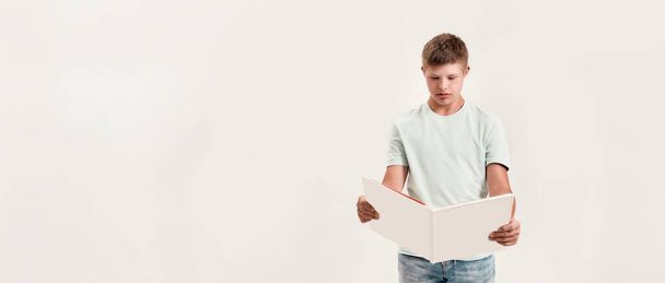 Teenaged disabled boy with Down syndrome looking focused while reading a book, standing isolated over white background - Zdjęcie, obraz