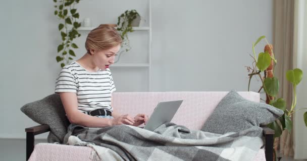 młody kaukaski student freelancer kobieta patrząc na komputer laptop czytanie dobre śmieszne wiadomości e-mail śmieje się toothy uśmiech spojrzenia na aparat siedzi na kanapie domu w salonie pokryte koc - Materiał filmowy, wideo