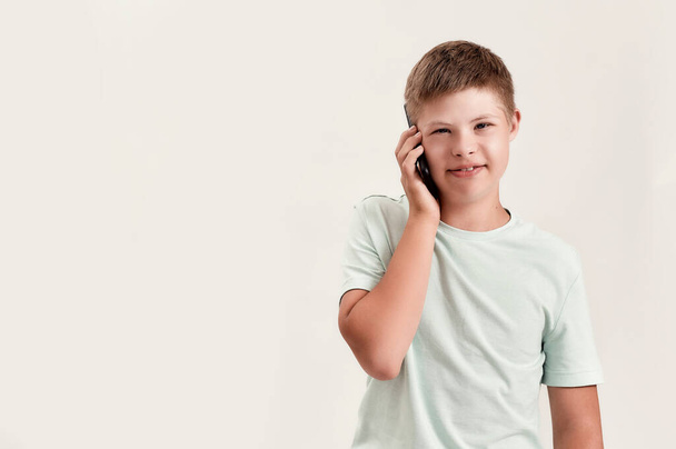 Portrait of teenaged disabled boy with Down syndrome smiling at camera while talking on the phone, standing isolated over white background - Foto, imagen