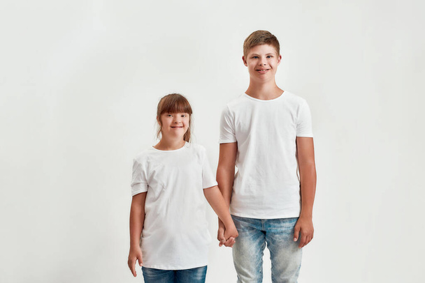 Two kids, disabled boy and girl with Down syndrome smiling at camera, holding each other hands while posing together isolated over white background - Fotó, kép