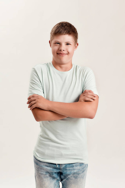 Teenaged disabled boy with Down syndrome smiling at camera while posing, standing with arms crossed isolated over white background - Foto, imagen