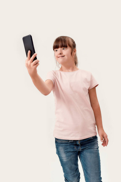 Portrait of disabled girl with Down syndrome using smartphone while taking selfie picture, standing isolated over white background - Fotoğraf, Görsel