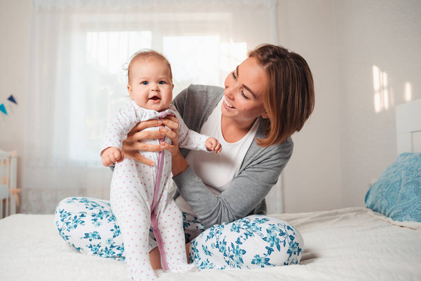 Joven madre feliz sosteniendo al bebé sonriente en sus manos mientras está sentada en la cama. Concepto de maternidad y cuidado infantil. - Foto, imagen