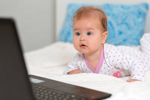 スライダーで驚くべき赤ちゃんの肖像画はベッドの上にあり、ラップトップを見ています。現代の技術に子供を教えるという概念. - 写真・画像
