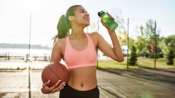 Happy young female basketball player smiling aside while drinking water from bottle, holding basketball, standing outdoors on a sunny day - Φωτογραφία, εικόνα