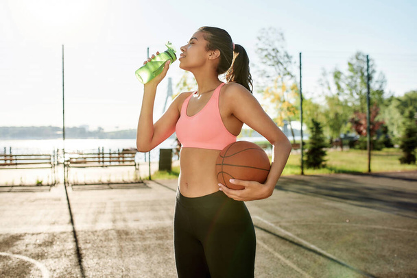 Attractive young female basketball player drinking water from bottle, holding basketball while standing outdoors on a sunny day - Φωτογραφία, εικόνα