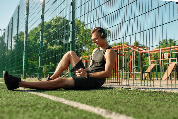 Young sportsman wearing headphones sitting on the field outdoors, listening to music, browsing playlist on smartphone and smiling - Φωτογραφία, εικόνα