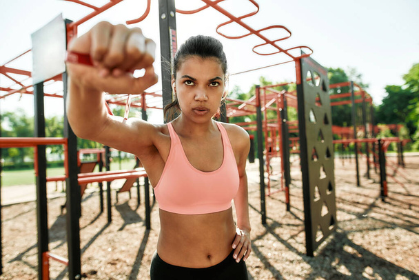 Portrait of concentrated young mixed race sportive woman having workout with rubber band at street gym yard - Foto, Imagen