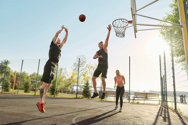 Athletic young friends spending time together outdoors on playground, playing basketball on a sunny day - Foto, afbeelding