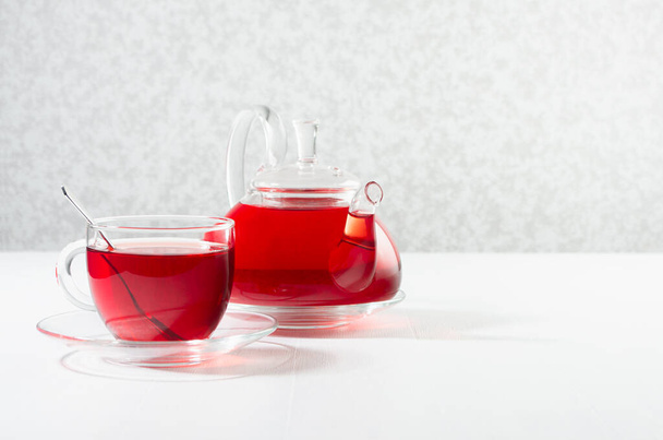 Red herbal tea in transparent tea pot with cup, spoon in sun beam in soft light white interior, copy space. - Фото, зображення