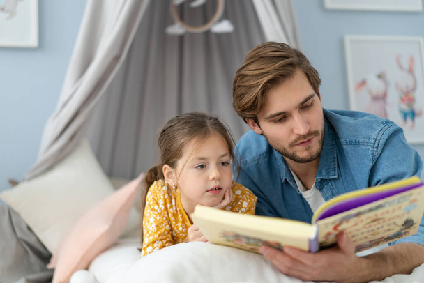 Father reading a book to his daughter while lying on the floor in bedroom. - Foto, Bild