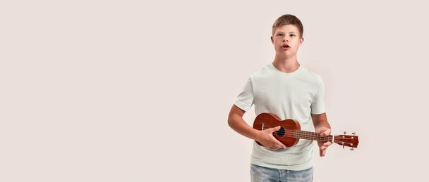 Teenaged disabled boy with Down syndrome holding, playing ukulele guitar, standing isolated over white background - Photo, image
