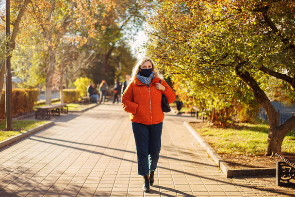 Sonbahar parkında güneşli bir günde salgın sırasında asfalt yolda yürüyen dış giysili ve maskeli kadın. - Fotoğraf, Görsel