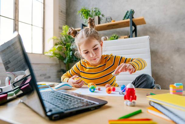 ラップトップを使用して8歳の少女は、部屋の机の上に座っている太陽系のオンライン惑星を研究する。若い女子小学生天文学のレッスンをオンラインで見て、彼女の宿題をする-子供粘土やプラスチックからの惑星モデルを彫刻 - 写真・画像