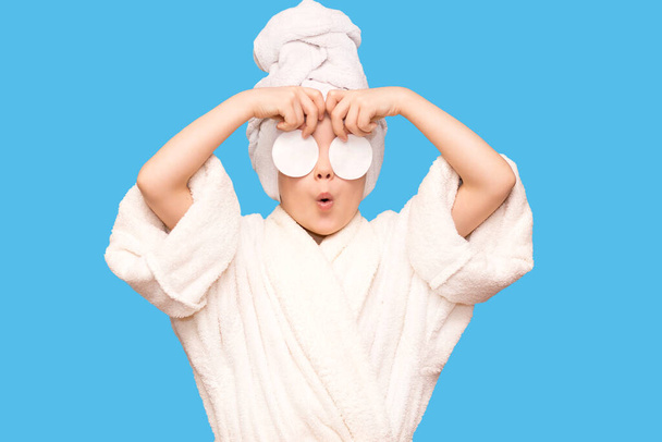 Beautiful kid girl with surprised face and towel on head hold cotton pad disk cleansing face skin with cleanser isolated on blue background. Happy girl remove makeup look at camera enjoy healthy clean skincare beauty treatment concept - Photo, Image