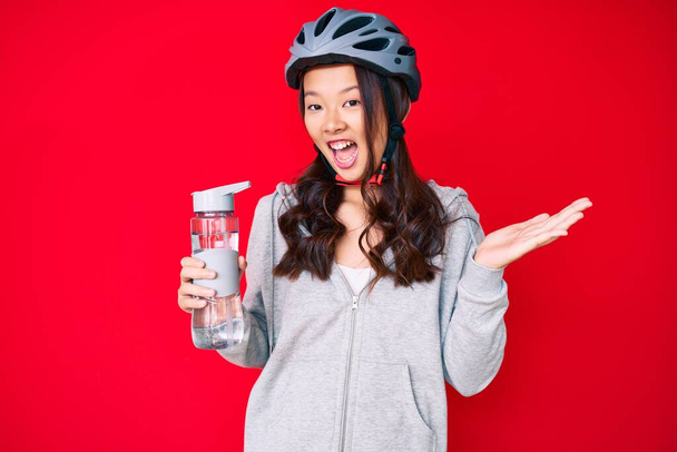 Menina chinesa bonita nova vestindo capacete de bicicleta e segurando garrafa de água celebrando realização com sorriso feliz e expressão vencedora com mão levantada  - Foto, Imagem