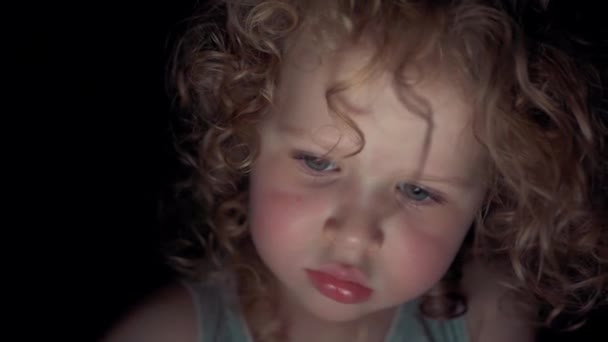 Κοντινό πρόσωπο ενός μικρού καυκάσιου σγουρού κοριτσιού που κοιτάζει την οθόνη μιας ψηφιακής συσκευής τη νύχτα. - Πλάνα, βίντεο