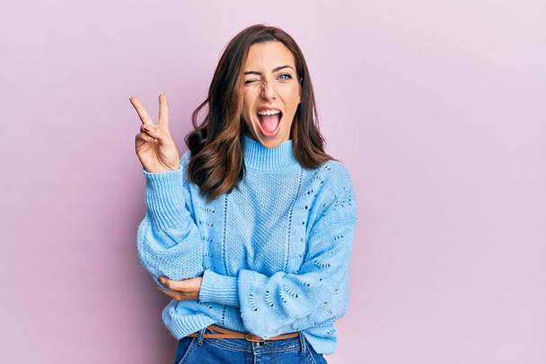 Young brunette woman wearing casual winter sweater over pink background smiling with happy face winking at the camera doing victory sign. number two.  - Photo, Image