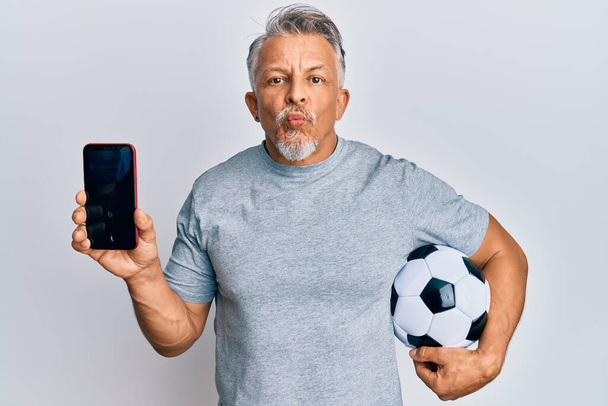 Mężczyzna w średnim wieku z siwymi włosami trzymający piłkę futbolową pokazującą smartfona patrzącego w kamerę dmuchającego pocałunek jest uroczy i seksowny. wyrażenie miłości.  - Zdjęcie, obraz