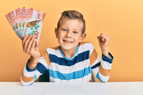 Очаровательный кавказский ребенок с банкнотой в 100 новых зеландских долларов сидит на столе и гордо кричит, празднуя победу и успех, очень взволнованный с поднятой рукой  - Фото, изображение