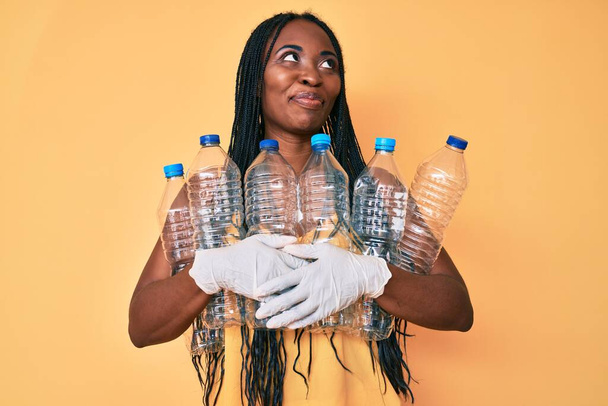 Saçları örgülü Afrikalı Amerikalı bir kadın plastik şişeleri geri dönüşüm kutusunu tutuyor gülümsüyor ve diğer tarafa bakıyor ve düşünüyor..  - Fotoğraf, Görsel