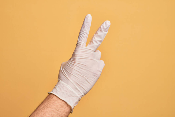 Käsi valkoihoinen nuori mies lääketieteellinen käsine yli eristetty keltainen tausta laskenta numero 2 osoittaa kaksi sormea, eleiden voitto ja voittaja symboli - Valokuva, kuva