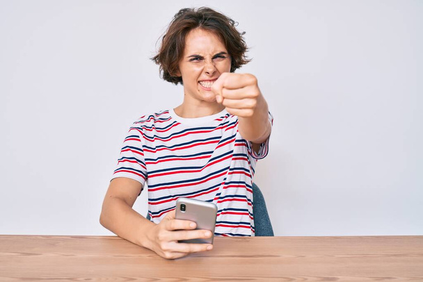 Mujer hispana joven usando teléfono inteligente sentado en la mesa molesto y frustrado gritando con ira, gritando loco con la ira y la mano levantada  - Foto, Imagen
