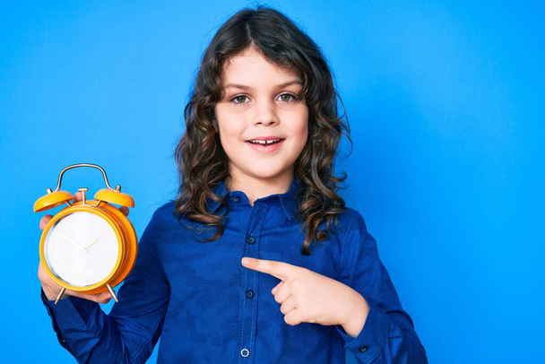 Cute Latynoski dziecko z długimi włosami trzymając budzik uśmiechnięty szczęśliwy wskazując ręką i palcem  - Zdjęcie, obraz