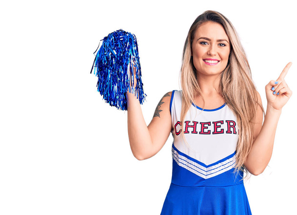 Young beautiful blonde woman wearing cheerleader uniform holding pompom surprised with an idea or question pointing finger with happy face, number one  - Photo, Image