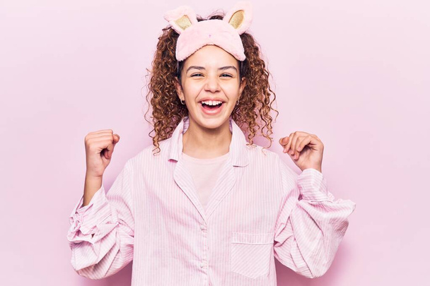 Beautiful kid girl with curly hair wearing sleep mask and pajamas screaming proud, celebrating victory and success very excited with raised arms  - Foto, imagen