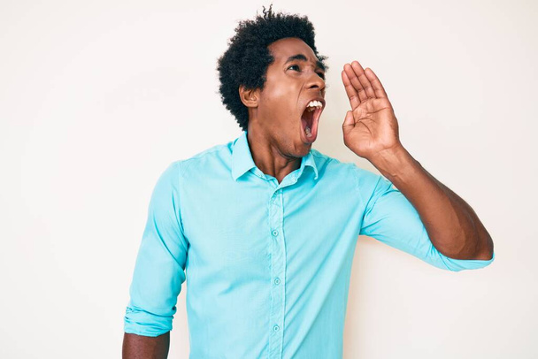 Bonito americano africano com cabelo afro vestindo roupas casuais gritando e gritando alto para o lado com a mão na boca. conceito de comunicação.  - Foto, Imagem