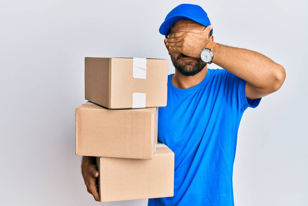 Handsome man with beard wearing courier uniform holding delivery packages covering eyes with hand, looking serious and sad. sightless, hiding and rejection concept  - Foto, Imagem
