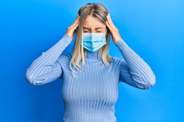 Beautiful blonde woman wearing covid-19 medical mask suffering from headache desperate and stressed because pain and migraine. hands on head.  - Photo, image