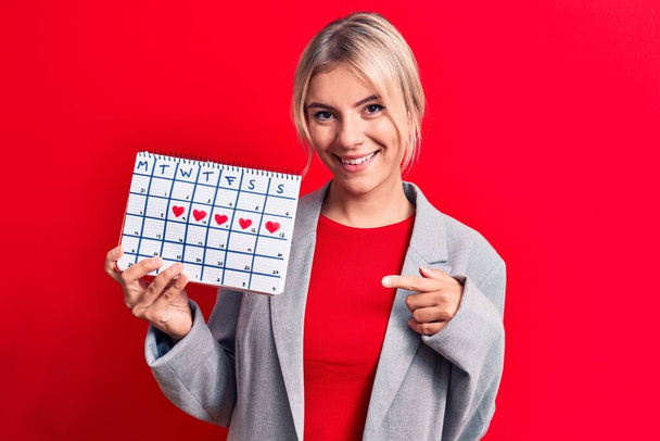schöne blonde Frau, die die Menstruation kontrolliert, hält Kalender über rotem Hintergrund und lächelt glücklich und zeigt mit Hand und Finger - Foto, Bild