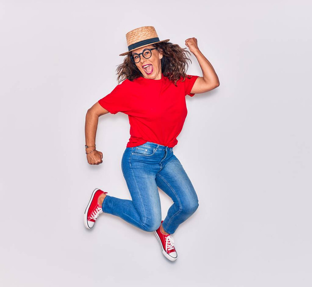 Mujer hispana hermosa de mediana edad vistiendo ropa casual y sombrero sonriendo feliz. Saltando con sonrisa en la cara sobre fondo blanco - Foto, imagen