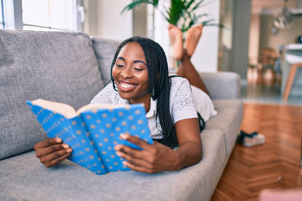 Νεαρή Αφροαμερικανή γυναίκα χαμογελά ευτυχισμένη ξαπλωμένη στον καναπέ διαβάζοντας το βιβλίο στο σπίτι - Φωτογραφία, εικόνα