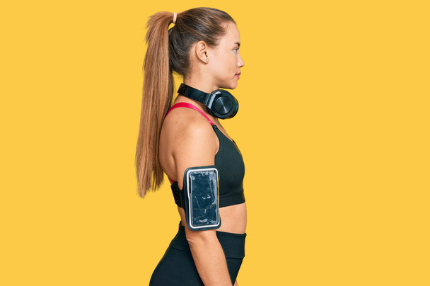 Beautiful blonde woman wearing gym clothes and using headphones looking to side, relax profile pose with natural face and confident smile.  - Foto, Imagem