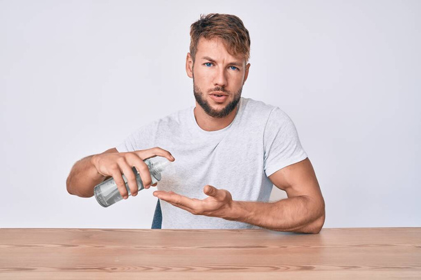 Jeune homme caucasien utilisant gel désinfectant pour les mains assis sur la table dans le visage de choc, l'air sceptique et sarcastique, surpris avec la bouche ouverte  - Photo, image
