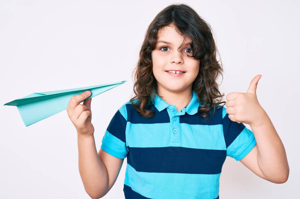 Мила іспаномовна дитина з довгим волоссям тримає паперовий літак усміхнений щасливий і позитивний, великий палець робить відмінний знак схвалення
  - Фото, зображення