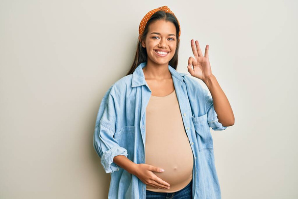 Mooie Latijns-Amerikaanse vrouw verwacht een baby, het aanraken van zwangere buik lachend positief doen ok teken met de hand en vingers. succesvolle uitdrukking.  - Foto, afbeelding