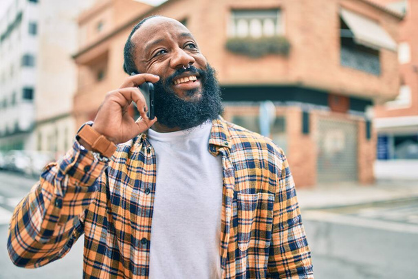 Jóképű, modern afro-amerikai férfi, szakállal a telefonban beszél a városban, boldog mosollyal. - Fotó, kép