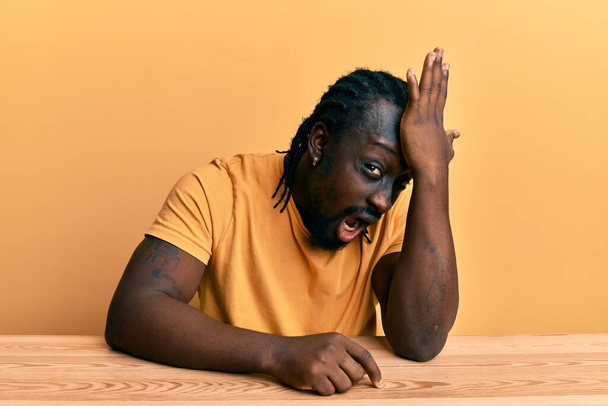Handsome young black man wearing casual clothes sitting on the table surprised with hand on head for mistake, remember error. forgot, bad memory concept.  - Фото, изображение